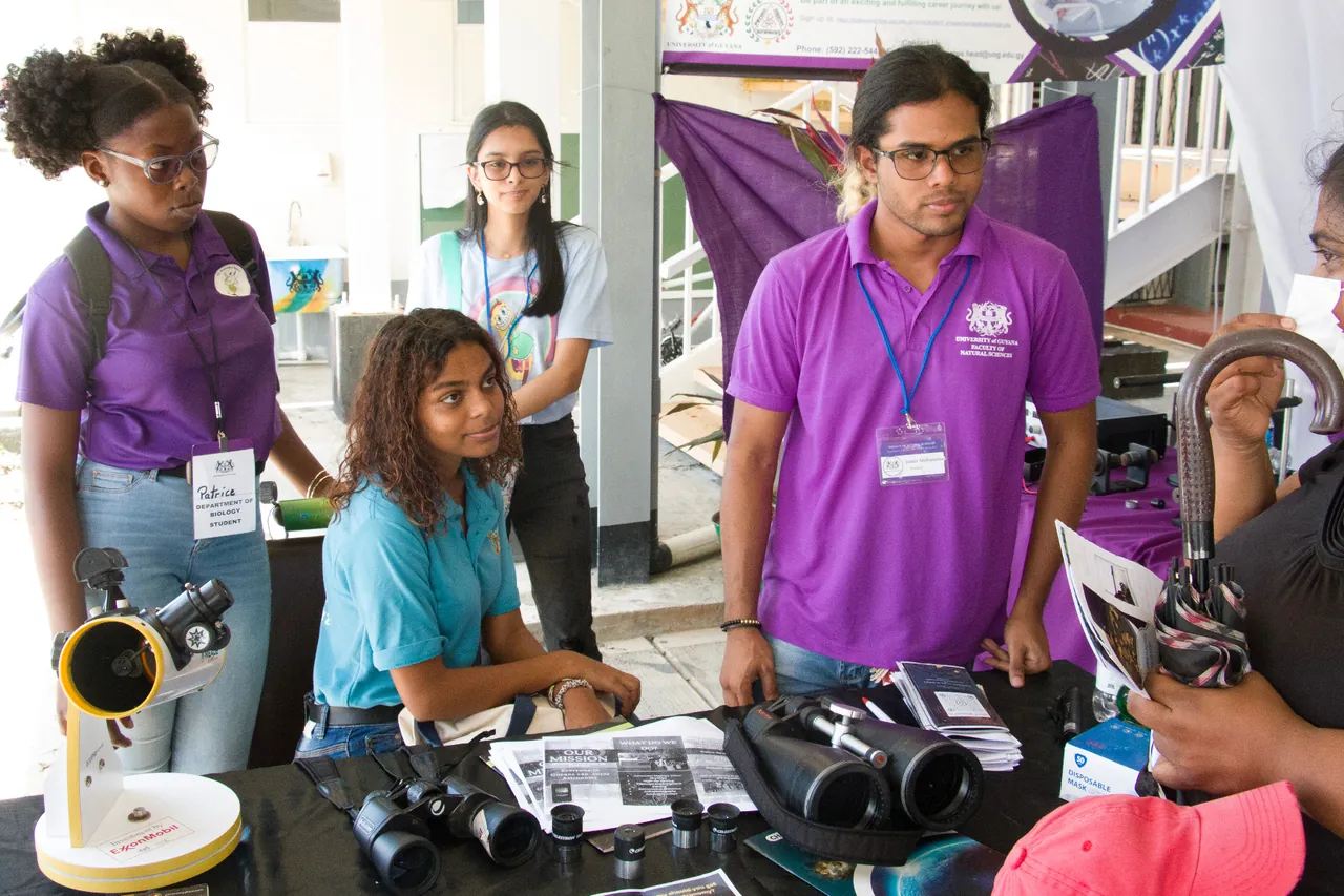 Samir with University of Guyana students at an outreach table laid out with binoculars, a small telescope and handouts